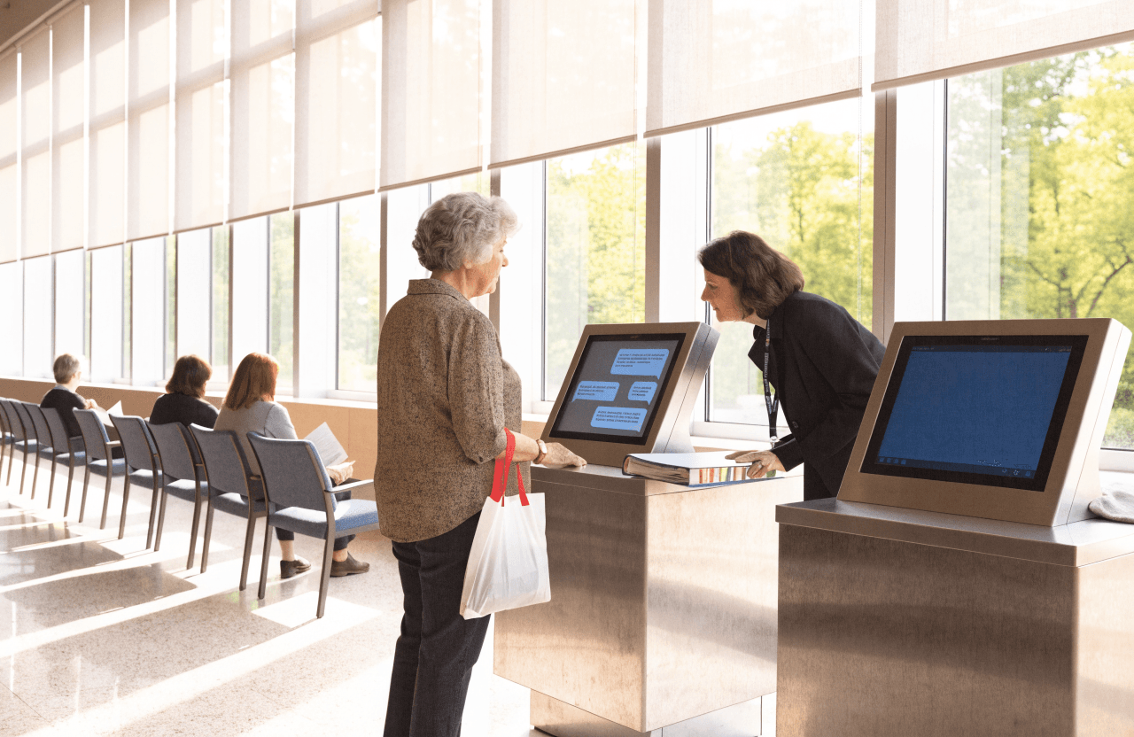 In a town hall's service hall, a woman asks a terminal a question while the city's AI assistant answers on screen and an official beside her reads along. A second terminal is switched off under a cloth.